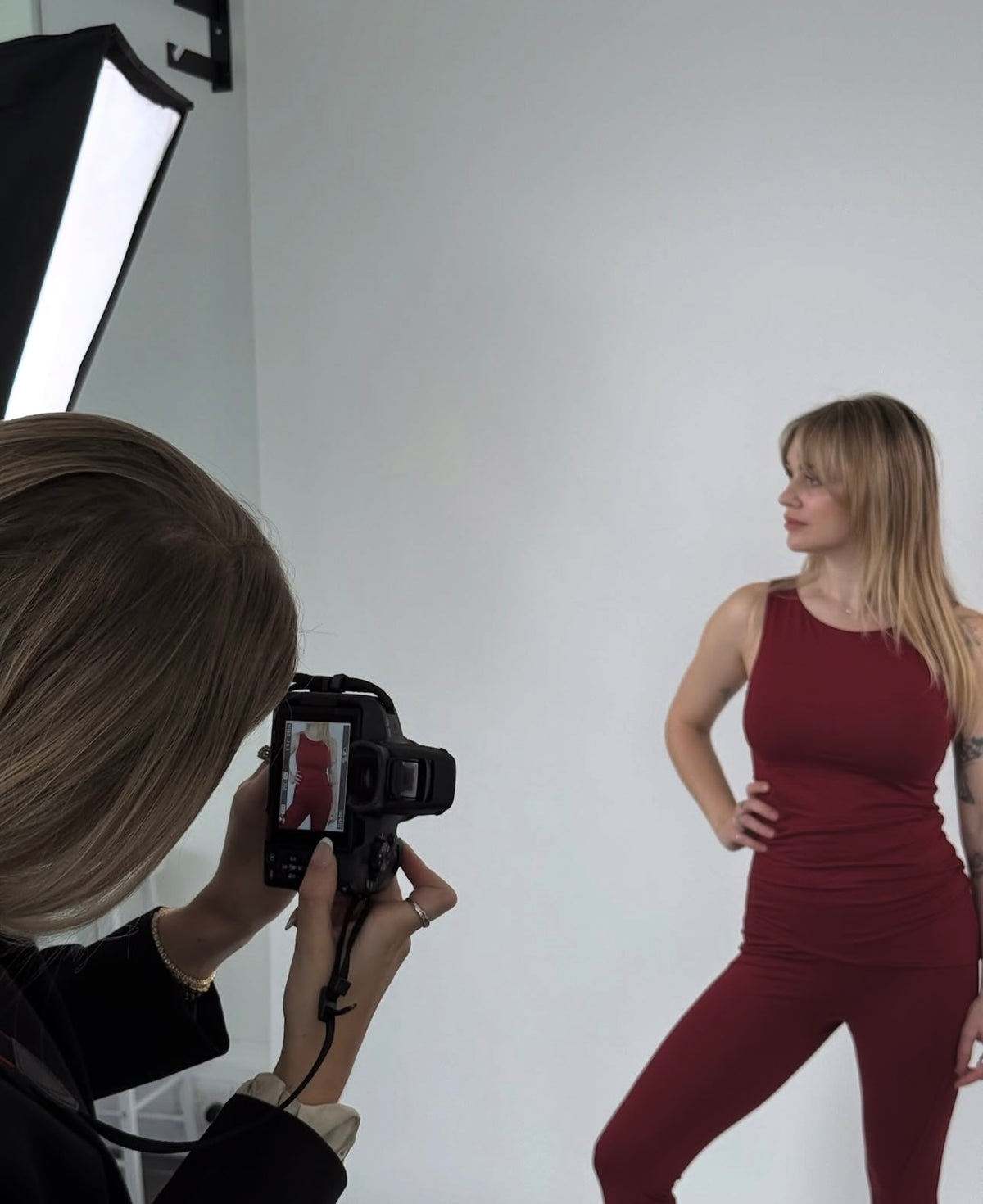 A model wearing a matching sleeveless top and leggings set in a deep burgundy red poses for a photo shoot, while a photographer in the foreground holds a DSLR camera. The background is a clean white studio backdrop with a softbox light visible on the left.