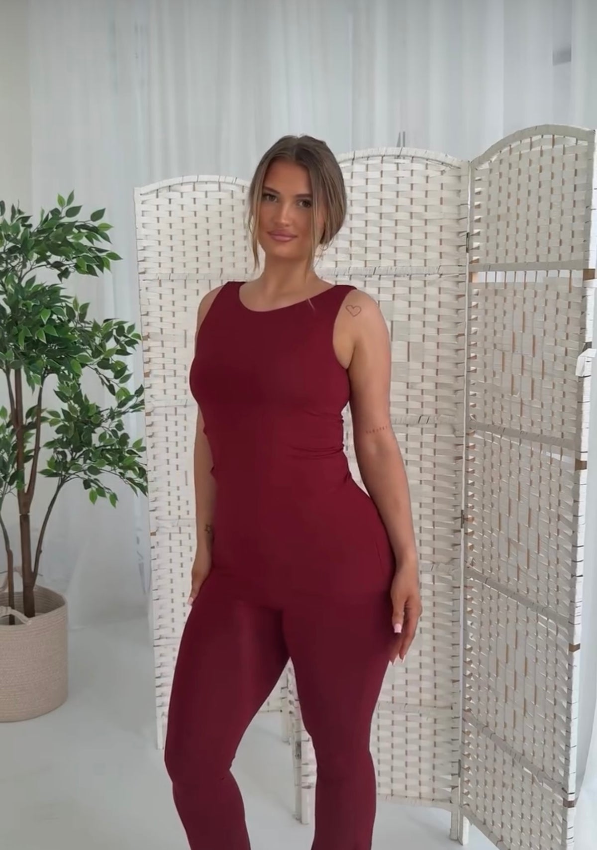 A woman modeling a sleeveless burgundy jumpsuit in a bright indoor setting with a white folding screen and a potted plant.