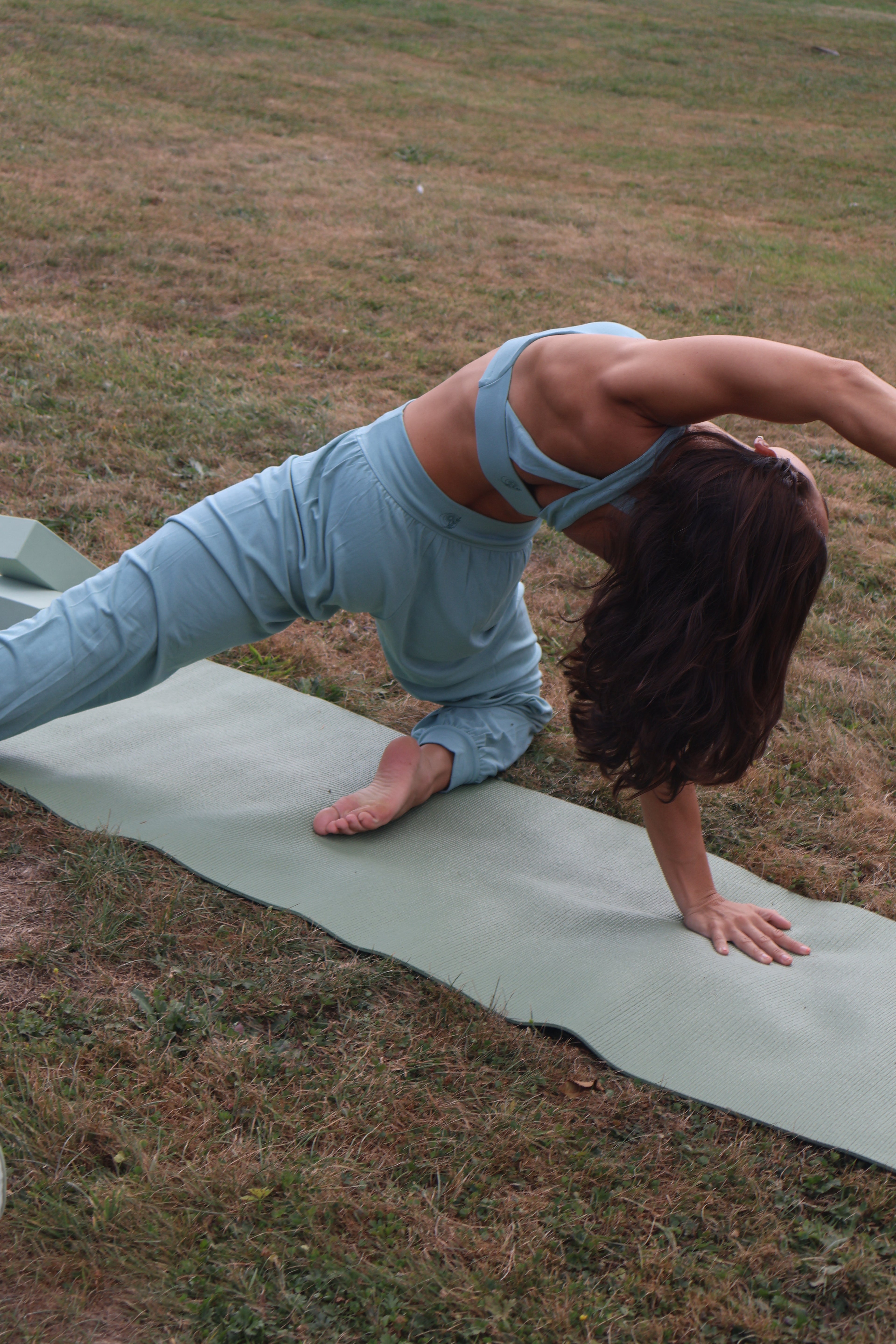 Woman in a sage green outfit stretching on yoga mat outdoors.